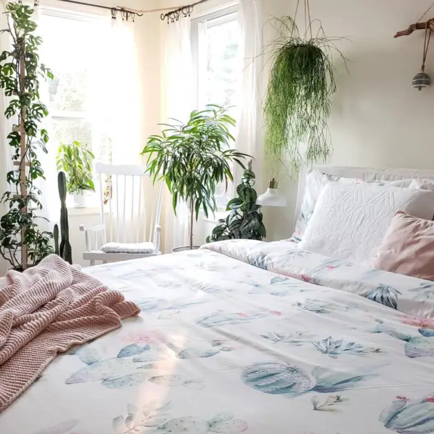 A sunlit bedroom featuring a bed with a whimsical cactus-print duvet, a soft pink throw, and numerous houseplants.