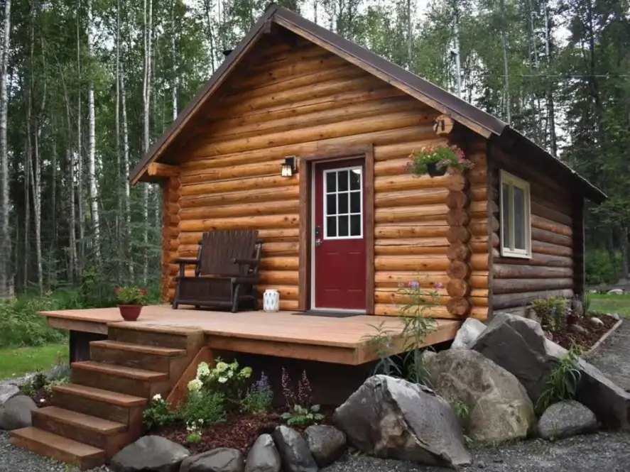 Small Log Cabins: 36 Ideas for Cozy Living & Enduring Charm 38 A small log cabin with a red door and a simple deck, landscaped with large natural boulders.