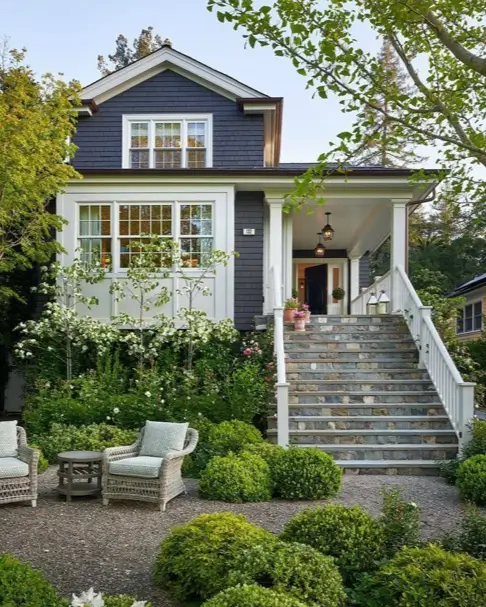 A sophisticated country house design featuring a grand stone staircase, dark shingle siding, and a terraced garden patio.