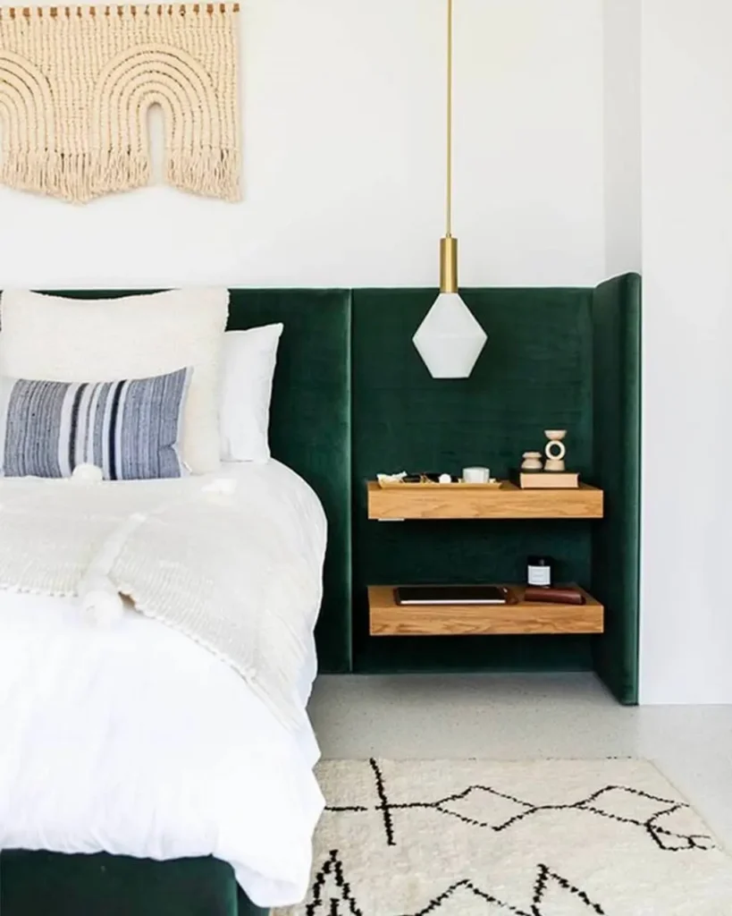 A modern bedroom with a large green velvet headboard, featuring a small, arched macrame wall hanging on the white wall above.