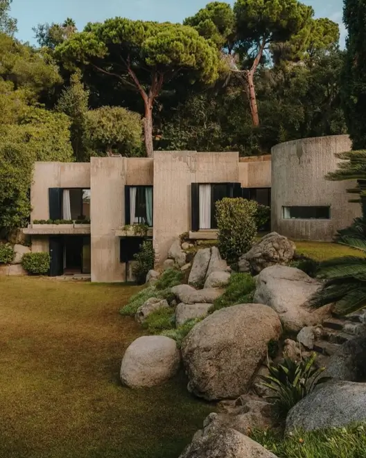 A brutalist home design with textured concrete walls and a prominent curved structure, settled into a landscape of large boulders and green lawn.