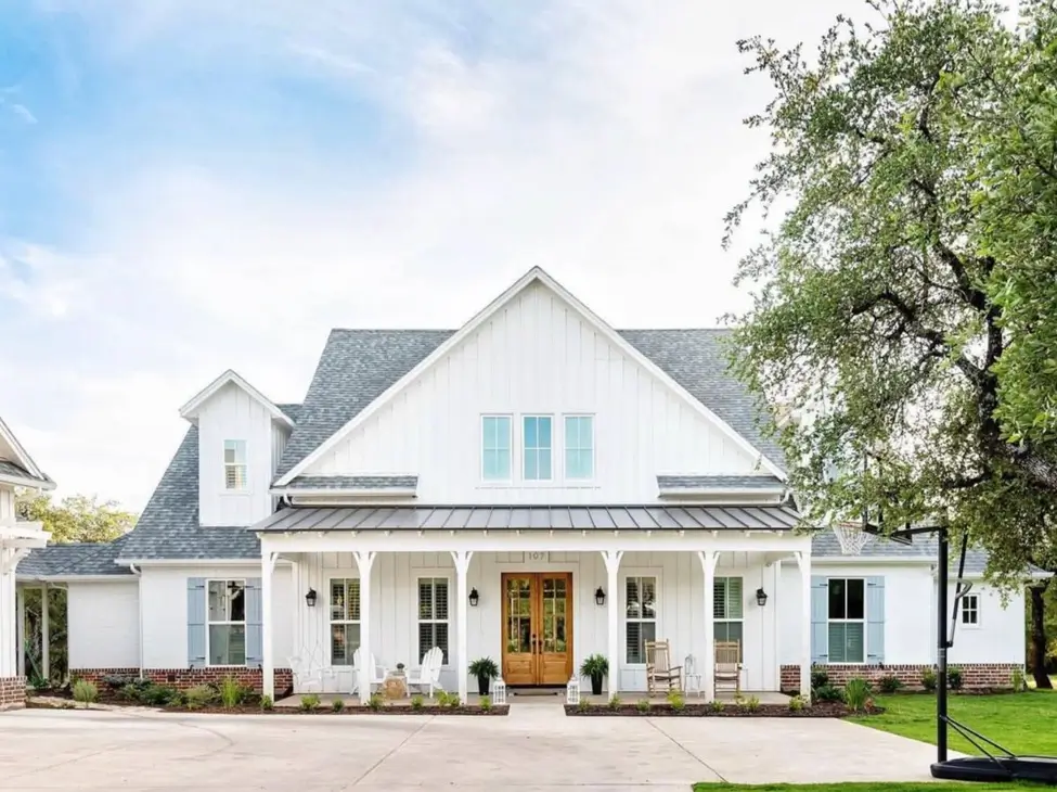 A welcoming off-white farmhouse exterior featuring a long front porch, light blue shutters, and a natural wood front door.