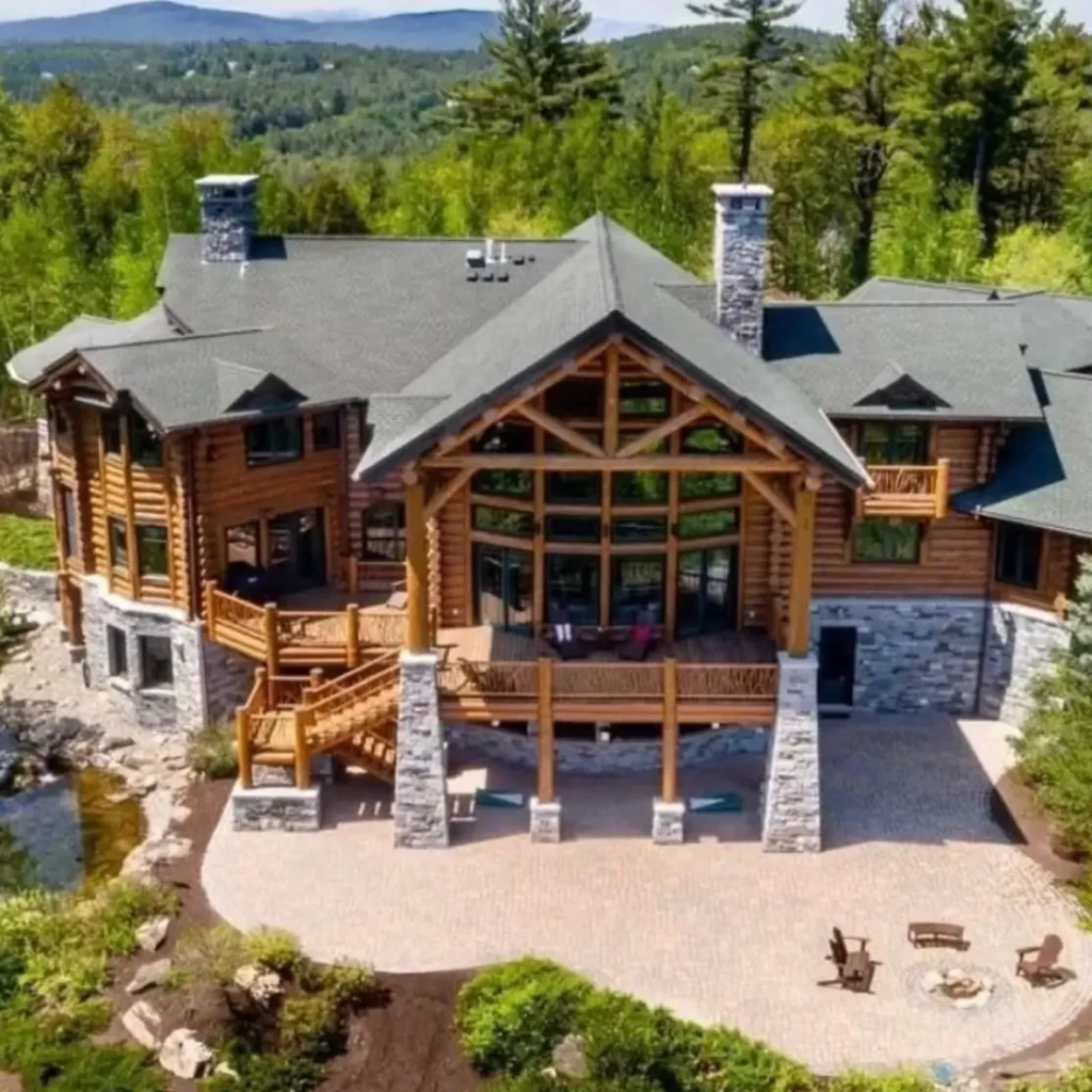 Embracing Grandeur: 25 Inspiring Ideas for Large Log Cabin Homes 7 An aerial view of a large log cabin home featuring a raised deck, a lower paver patio, and stone accents overlooking a forest.