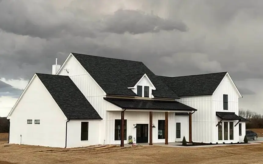 A modern white farmhouse exterior with vertical siding and multiple gables under a dramatic, cloudy sky, featuring black window frames and a small porch with wood posts.