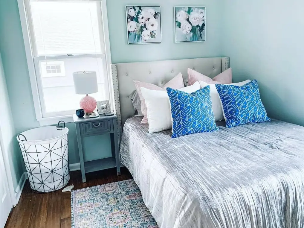 A small cozy bedroom idea with light blue walls, a tufted headboard, and bold blue and pink accent pillows on the bed.