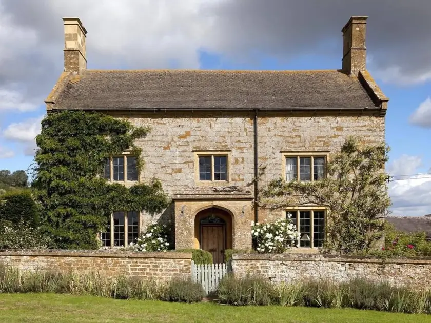 A symmetrical stone British country house with prominent twin chimneys and climbing plants behind a low stone wall.