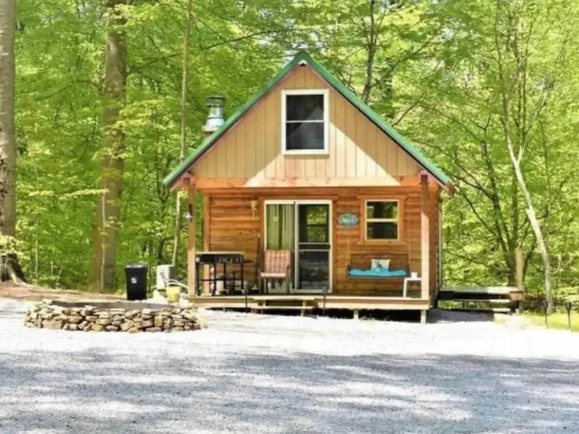 60 Charming Small Log Cabin Ideas to Inspire Your Rustic Retreat 44 A small wooden cabin with a green roof and a stone fire pit in a gravel yard, surrounded by green trees.