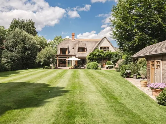 A country house design with a thatched roof overlooking a vast, perfectly striped lawn from the back patio.
