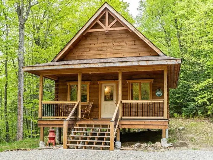 60 Charming Small Log Cabin Ideas to Inspire Your Rustic Retreat 46 An elevated small log cabin on piers with a covered front porch and rocking chairs, surrounded by lush green forest.