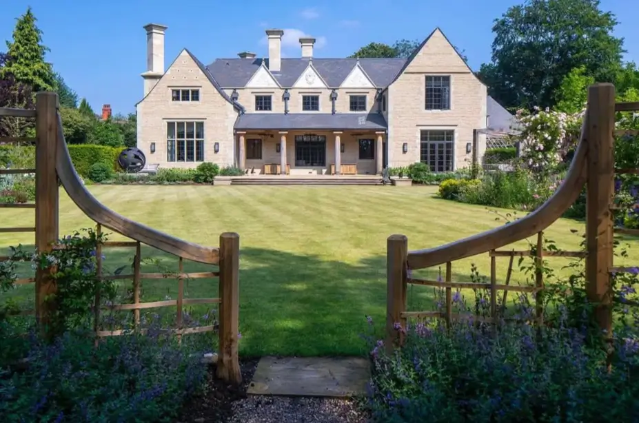 A modern stone country house design viewed through a beautifully curved wooden garden trellis framing the expansive back lawn.