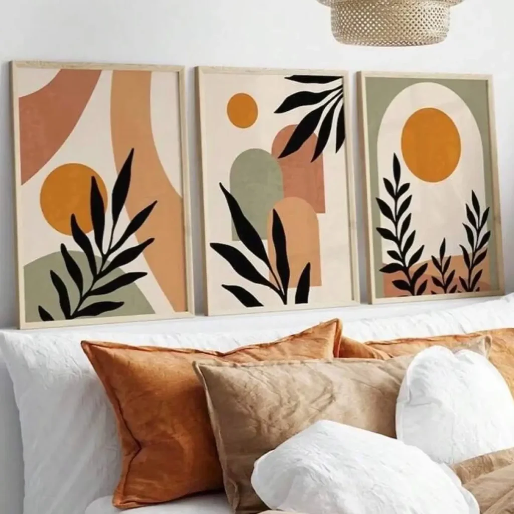 A set of three matching abstract art prints in light wood frames, featuring sun and leaf motifs, hanging over a bed with rust and white pillows.