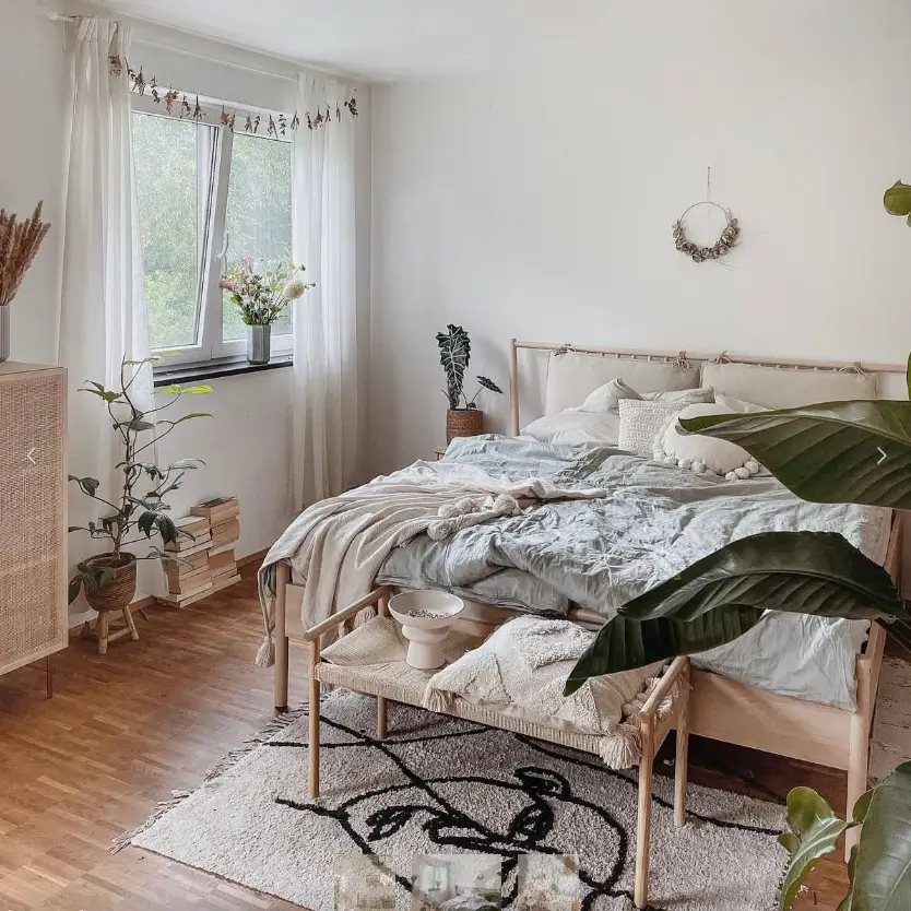 A bright bedroom with a light wood bed dressed in soft, rumpled blue-gray linens and surrounded by plants.