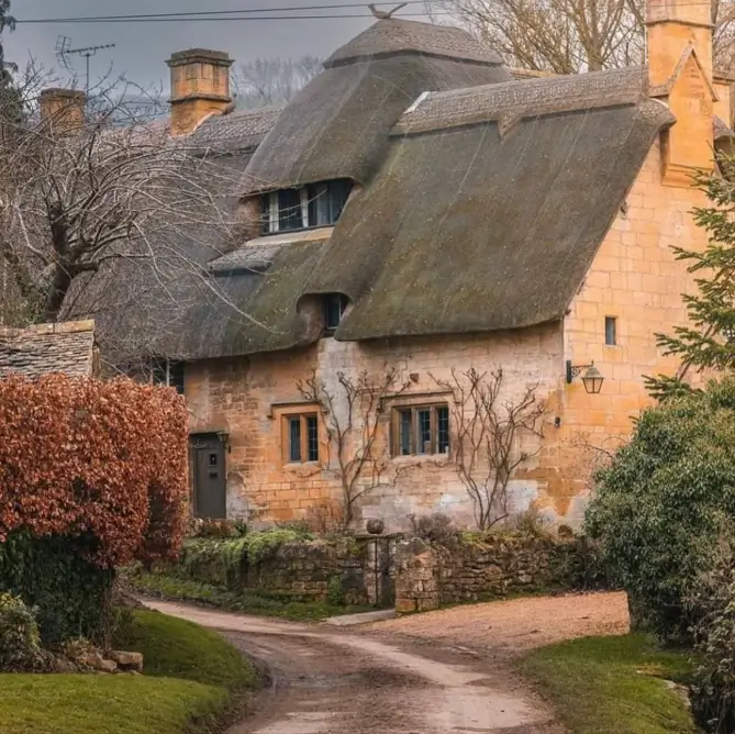 Quaint English country cottage with a sculptural thatched roof and stone walls on a winding country lane.