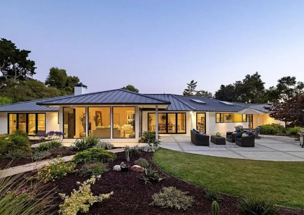 A modern white ranch house with a large back patio and glass walls illuminated from within at twilight.