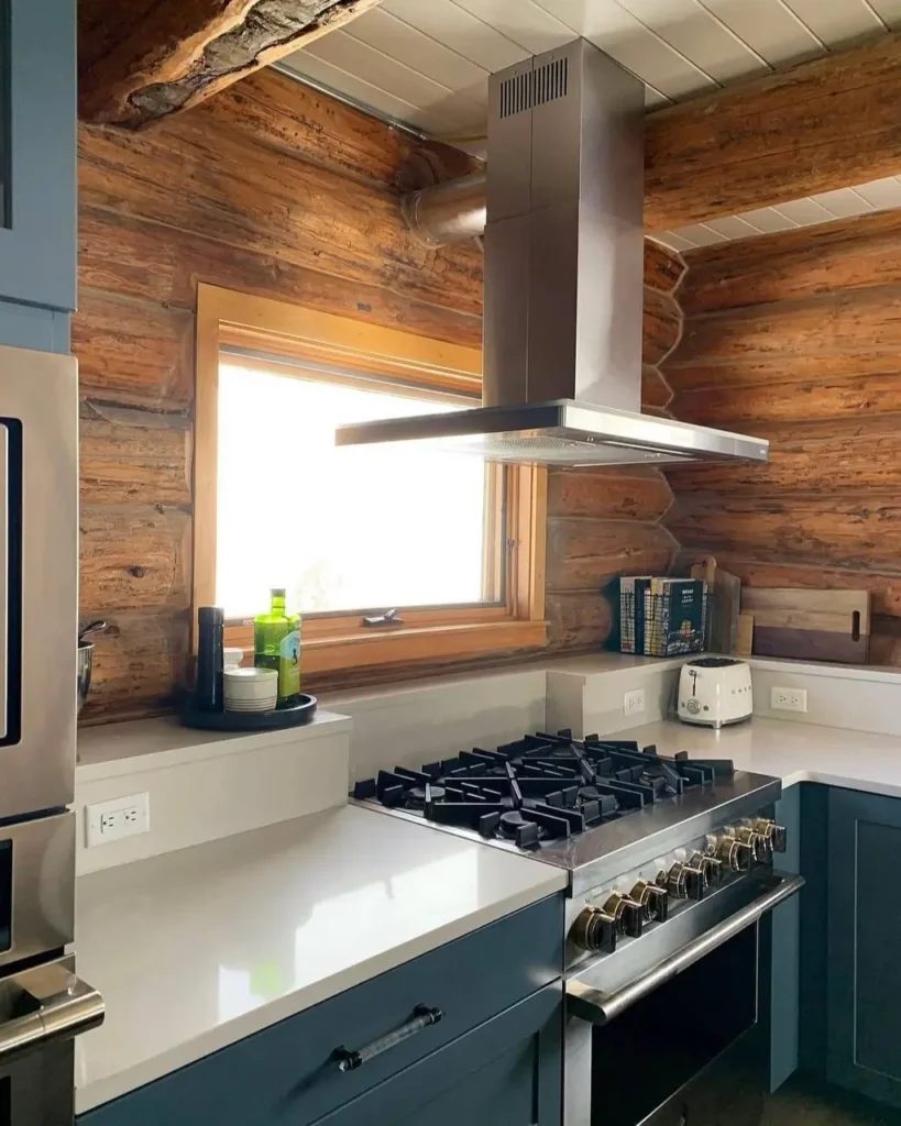 A modern rustic kitchen featuring traditional log walls contrasted with a sleek stainless steel range hood and dark blue cabinets.