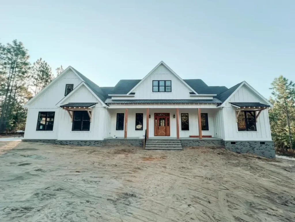 A symmetrical modern white farmhouse exterior with a central wood front door, black windows, and a gray brick foundation skirt, set on a dirt lot with pine trees behind.