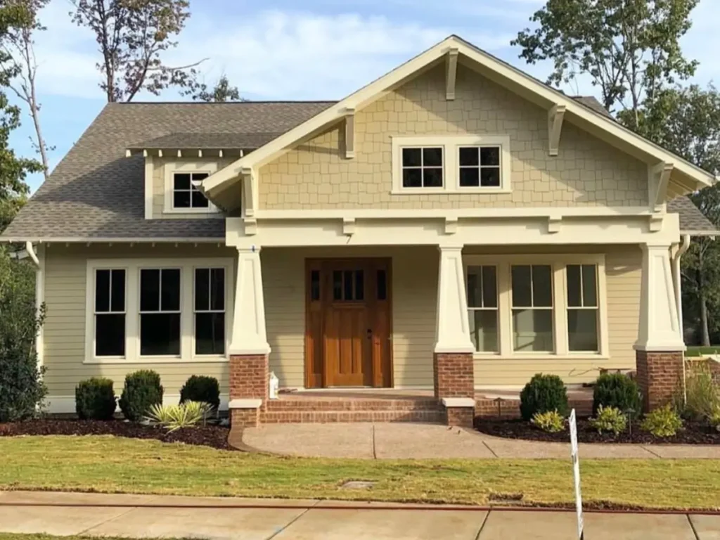 A single-story craftsman house exterior with warm tan siding, a front porch with brick piers, and a natural wood door.
