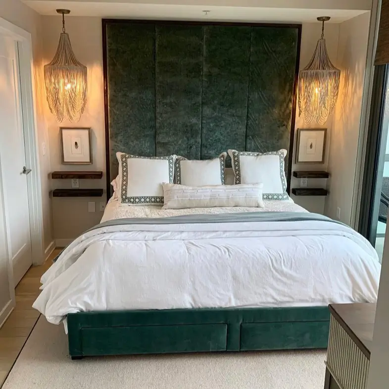 A small, luxurious bedroom interior design with a tall green velvet headboard and two dramatic beaded chandeliers hanging over floating nightstands.