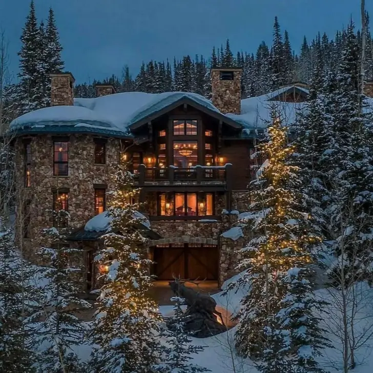 16 Mountain Mansions That Define Luxury Alpine Living 9 Magical winter scene of a snow-covered stone mountain mansion, its cozy interior glowing alongside festively lit pine trees.