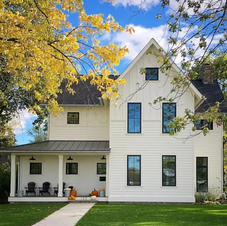 A two-story off-white farmhouse with black windows, framed by trees with bright yellow autumn leaves.