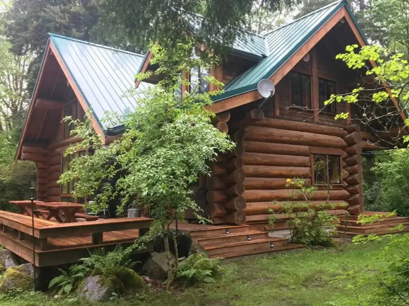 60 Charming Small Log Cabin Ideas to Inspire Your Rustic Retreat 54 A small log cabin with a multi-gable green metal roof and a large wooden deck, surrounded by forest.
