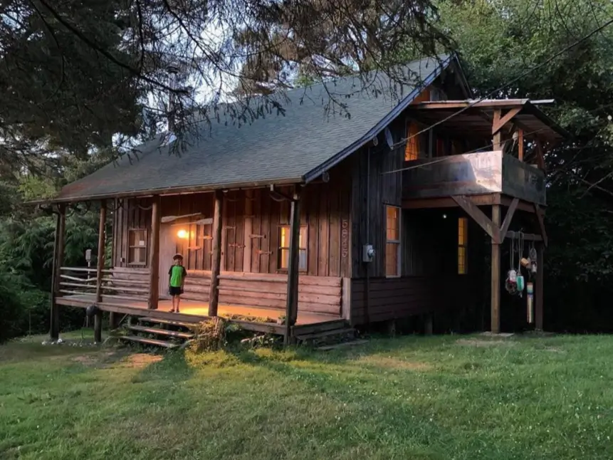 60 Charming Small Log Cabin Ideas to Inspire Your Rustic Retreat 57 A rustic wooden cabin at dusk with a wraparound porch and an elevated covered balcony.