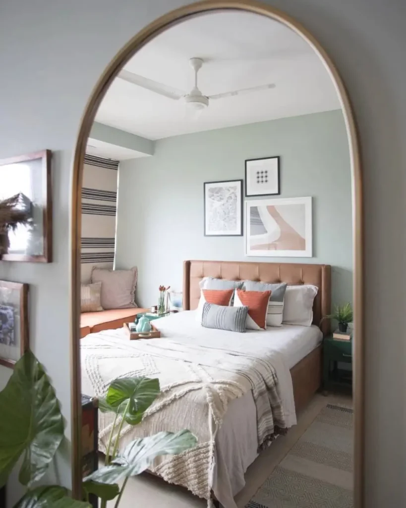 A reflection in an arched mirror showing a bedroom with a gallery wall of three abstract prints above a brown leather headboard.