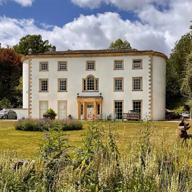 A large white British country house featuring a distinctive curved front facade and a sprawling lawn.