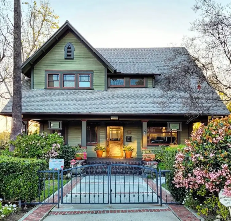 A historic craftsman house exterior featuring sage green siding, dark olive trim, and a prominent front gable surrounded by mature landscaping.
