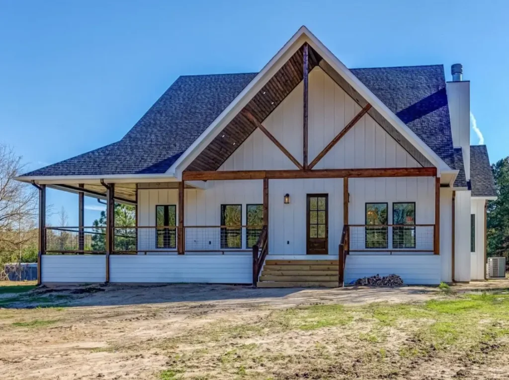 The rear view of a modern white farmhouse featuring a large gable with a dark wood decorative truss, a wraparound porch, and modern cable railings.