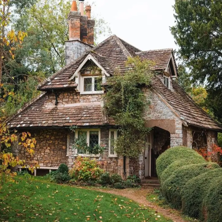 A rustic stone cottage with an asymmetrical roof and climbing plants nestled among trees.