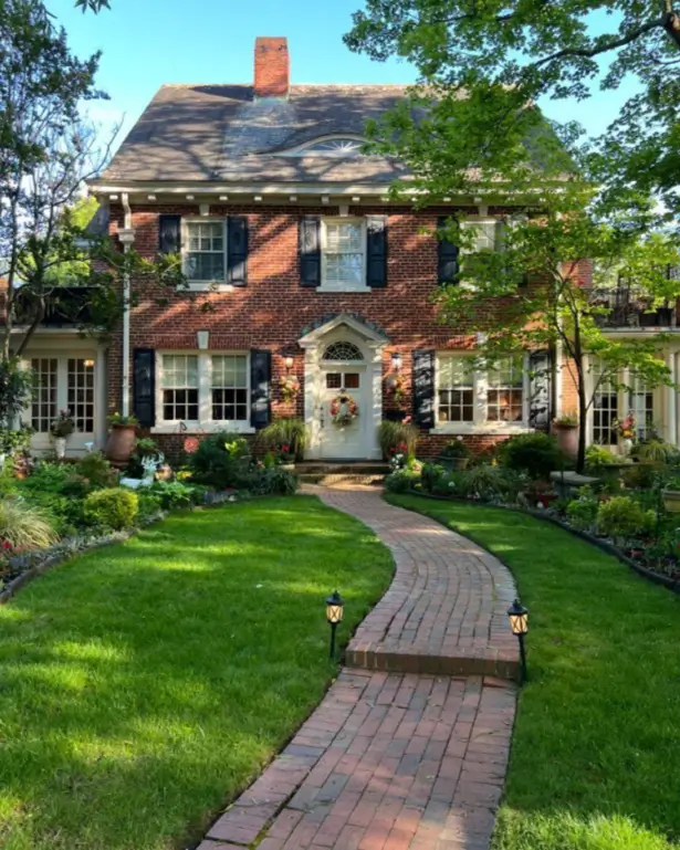 A stately red brick country house design with black shutters, a curving brick path, and formal gardens.