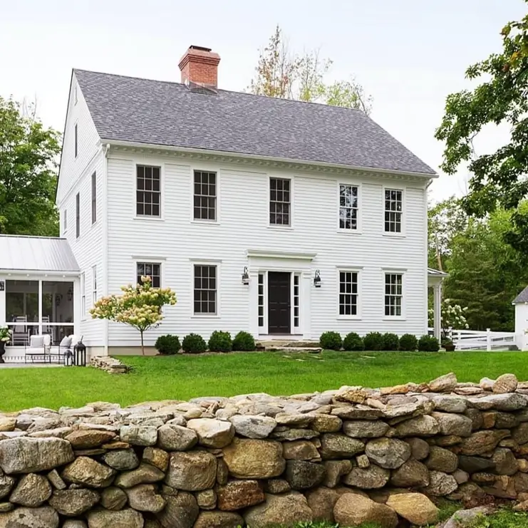 A traditional off-white colonial farmhouse with a prominent red brick chimney and a rustic stone wall in the yard.