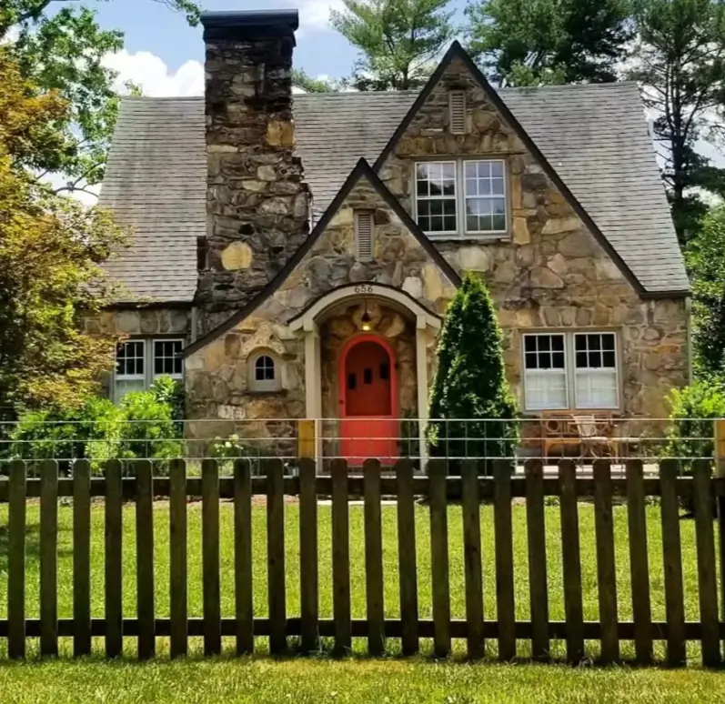 A stone cottage house design with a large stone chimney and a bright red arched door.