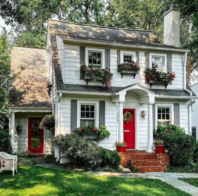 A charming white country house design featuring gray shutters, vibrant red doors, and overflowing flower window boxes.