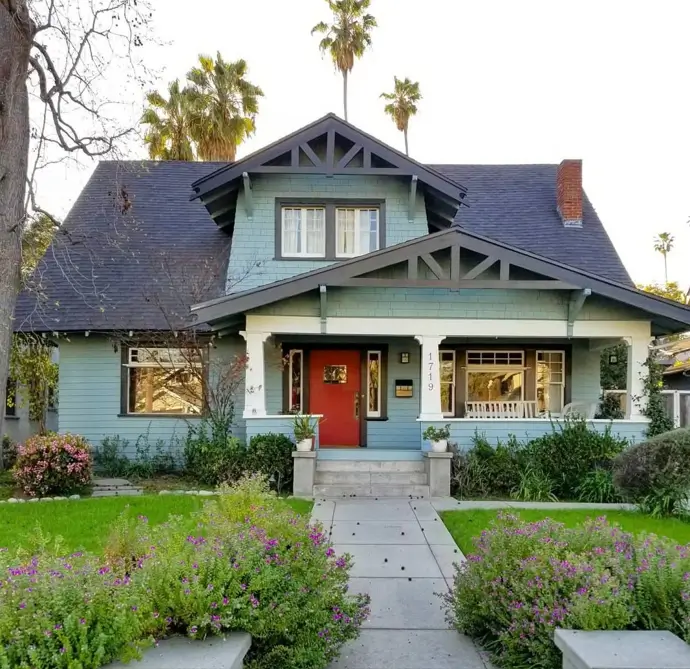 A craftsman house exterior in a soft blue-green color with dark exposed trusses, a white porch, and a bold red front door.