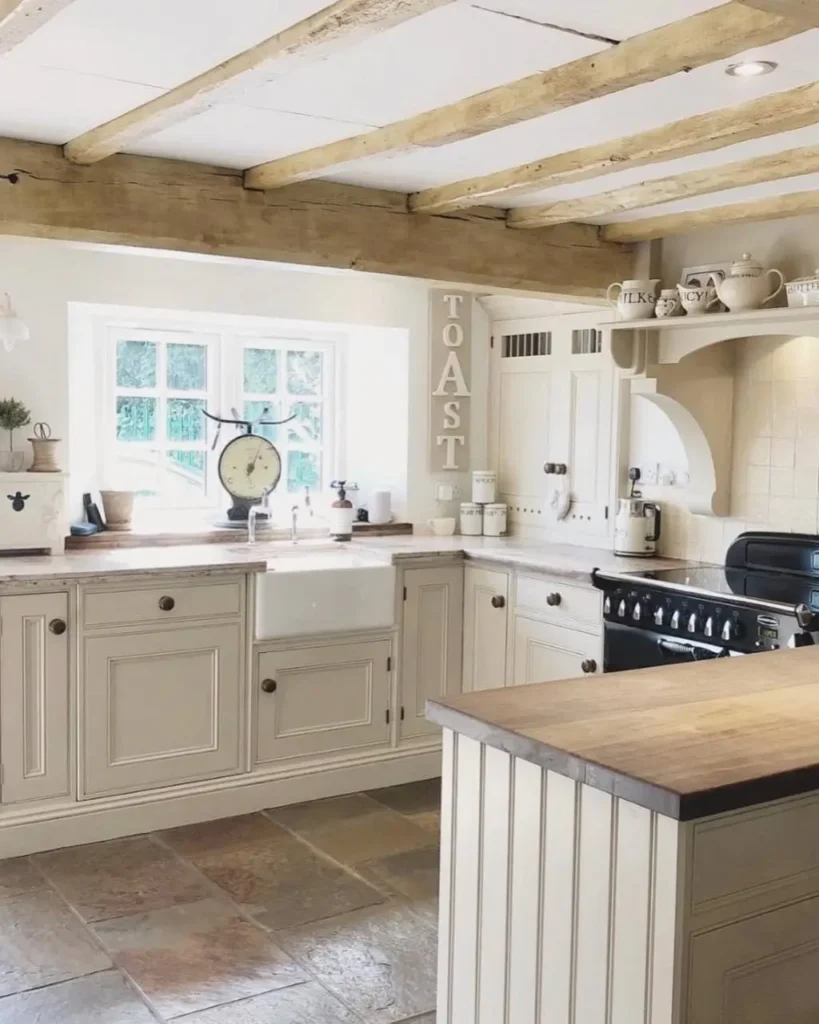 A light and airy rustic kitchen design with creamy white cabinets, exposed wood beams, and a central farmhouse sink.