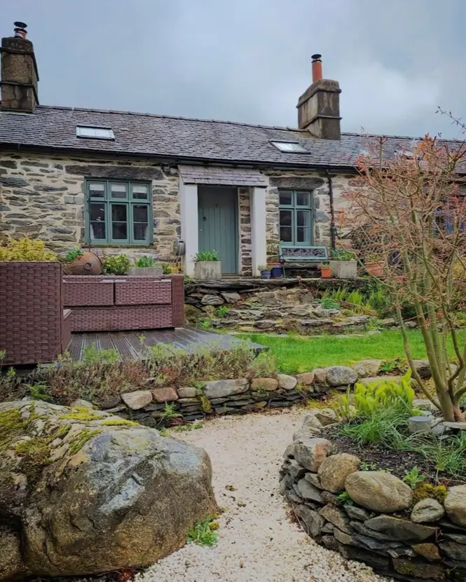 A long, single-story stone cottage with green trim, a patio, and a terraced garden.