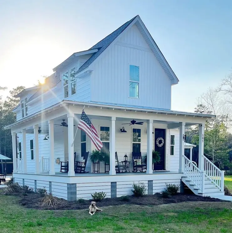 A sunny off-white farmhouse with a large wraparound porch furnished with rocking chairs and an American flag.