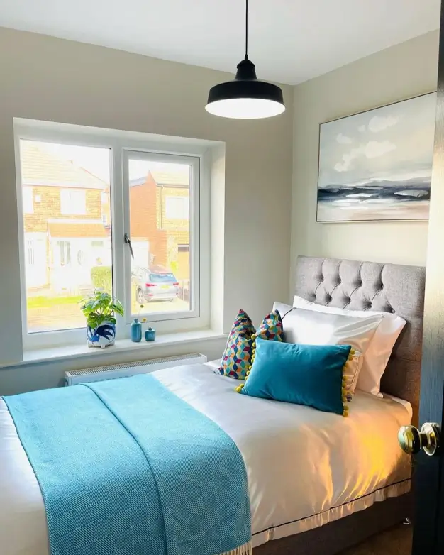 A small cozy bedroom design with a grey tufted headboard, white linens, and a bright turquoise throw blanket and accent pillow.