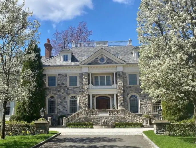 20 Stone Mansion Ideas for Classic & Modern Designs 10 A grand stone mansion with a dual staircase leading to a covered porch, framed by white blossoming trees under a blue sky.