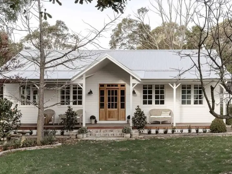 A charming off-white farmhouse cottage with a wooden deck porch and a light gray metal roof, nestled in a wooded setting.