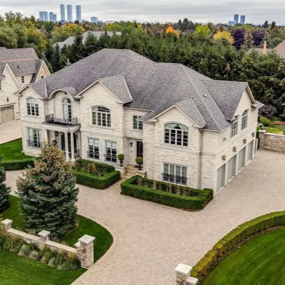 20 Stone Mansion Ideas for Classic & Modern Designs 11 An aerial view of a large, light-colored modern stone mansion with a paver driveway, manicured hedges, and a city skyline in the distance.