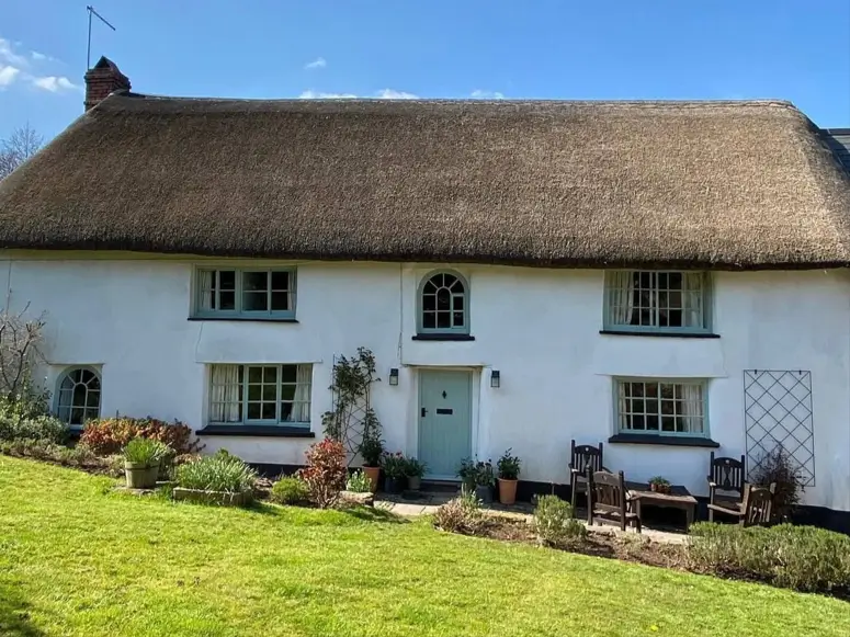 A white thatched English country cottage with light green trim and a central arched window above the door.