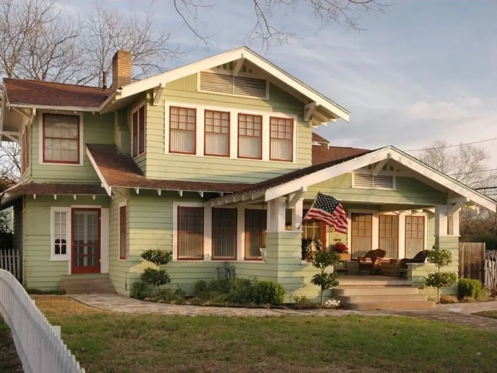 A classic American craftsman house with light green siding, contrasting red window trim, and a wide, welcoming front porch.