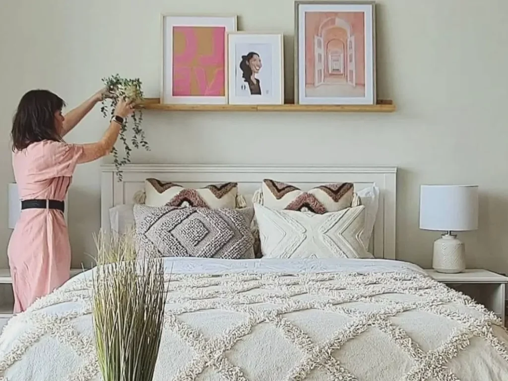 A woman in a pink dress styling a wooden picture ledge with framed boho wall art and a plant above a bed with textured white bedding.