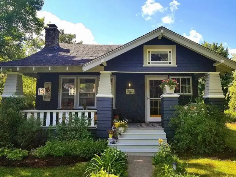 A cozy craftsman bungalow with deep navy siding, creamy off-white trim, and a rustic stone chimney.