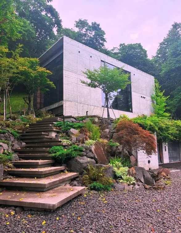 A contemporary brutalist house with a clean concrete form and large glass windows, set into a lush, rocky hillside with a floating concrete staircase.