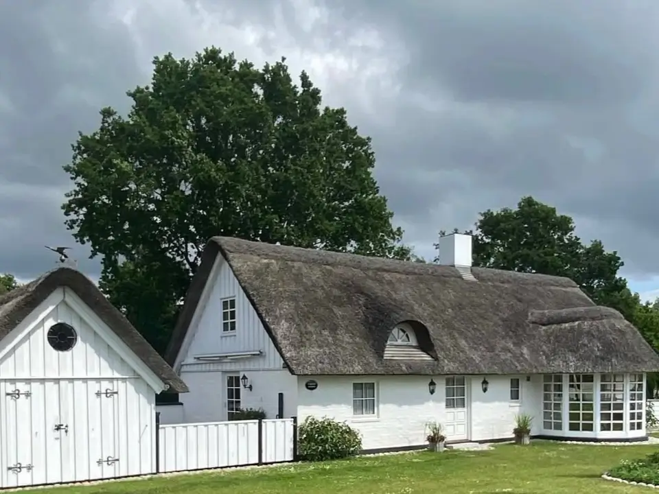 A classic country house design featuring a charming white cottage with a traditional thatched roof and a matching barn under a cloudy sky.