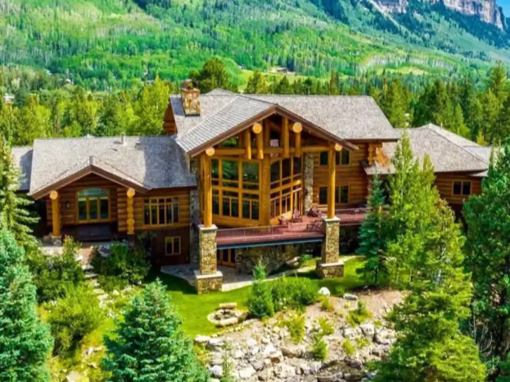 A grand and large log cabin home with stone pillars and an expansive deck, nestled in a lush green mountain forest.